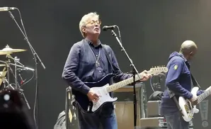 Guitarist Eric Clapton sings and plays a black Fender Stratocaster on stage with a bassist.