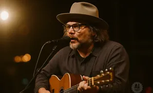 A man in a hat and glasses plays an acoustic guitar and sings into a microphone on stage.