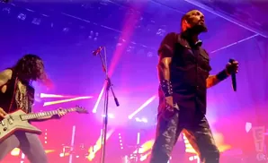 A male rock singer with a microphone stands center stage in front of purple and pink lights, while a guitarist plays to his left.