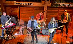 A band plays on stage, with a saxophonist/keyboardist, guitarist, drummer, and two bassists.