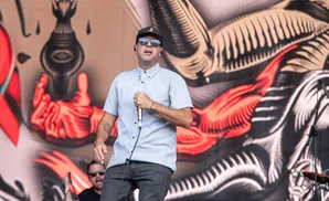 A man in a hat and sunglasses sings into a microphone on stage.