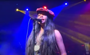 Singer with long dark hair and a red tartan hat performs on stage with blue and yellow spotlights.