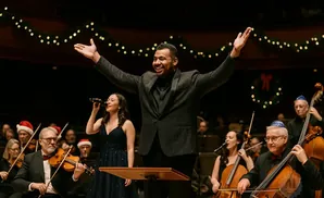 A conductor with arms raised smiles at a Christmas concert with an orchestra and singers.