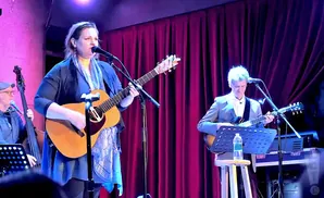 A woman plays guitar and sings into a microphone at a concert, with two men playing instruments behind her.