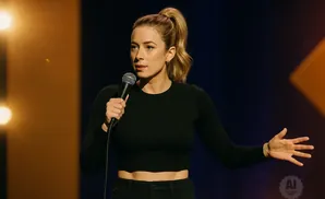 A woman with blonde hair in a ponytail, wearing a black crop top, holds a microphone and gestures with her open hand on stage.