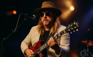 Musician in a hat and sunglasses plays a red electric guitar on stage.