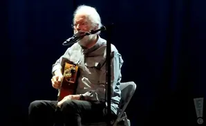 An older man with white hair and beard plays an acoustic guitar on stage.
