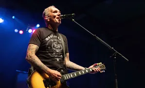 A tattooed man in a black t-shirt plays a guitar and sings into a microphone on a stage.