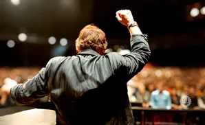 A man in a black jacket raises his fist to a cheering crowd from a stage.