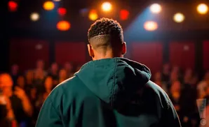 A man with a fade haircut stands with his back to the camera, wearing a teal hoodie on a dimly lit stage with a blurred audience.