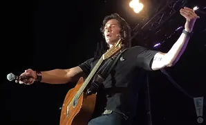 A male singer with long dark hair holds a microphone and plays an acoustic guitar on stage, wearing a black shirt and jeans.