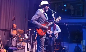 A man in a hat and suit plays an electric guitar on stage with a drummer and bassist.