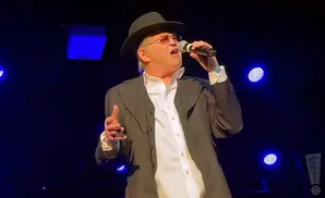 Singer in a black hat and suit, holding a microphone and gesturing with his hand, with blue stage lights in the background.
