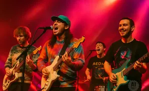 A band performs on stage with red lighting, four members playing guitars and singing.