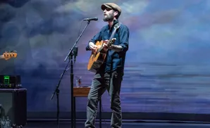 A man in a cap plays an acoustic guitar on stage with a blue, cloudy backdrop.