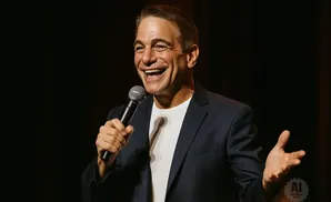 A man in a dark suit and white t-shirt smiles and holds a microphone.