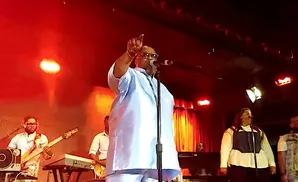A male singer in a light blue outfit stands center stage with a microphone, pointing up at a concert.
