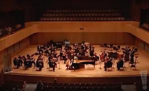 An orchestra performs on a stage with a grand piano at the center.