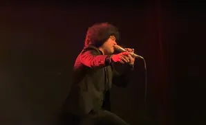 A man with an afro sings into a microphone on stage, wearing a black suit and red-lit jacket.