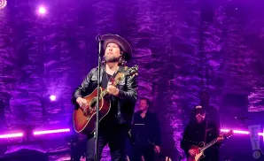 A man in a cowboy hat and leather jacket plays an acoustic guitar on stage with purple lighting.