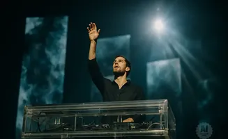 A DJ raises his hand in a dark, moody concert hall as spotlights shine down.
