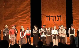 A group of actors in traditional clothing stand on stage in front of an orange backdrop with Hebrew text.