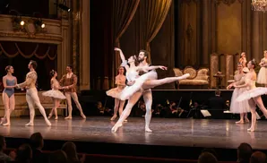 Dancers in elaborate costumes perform on a stage with dramatic lighting and ornate theatre boxes.