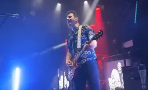 A male guitarist in a patterned shirt plays a red electric guitar on a brightly lit stage.