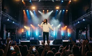 A male singer in a beige hoodie performs on a stage with blue and orange lights, while a crowd cheers below.