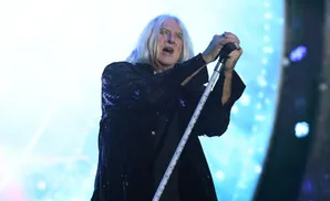 A man with long, white hair sings into a microphone, wearing a sequined jacket.