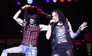 Two male rock musicians performing on stage, one holding a guitar above his head and the other singing into a microphone.