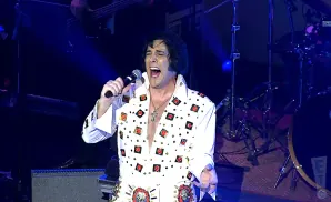 An Elvis impersonator in a white sequined jumpsuit sings into a microphone on a dimly lit stage.
