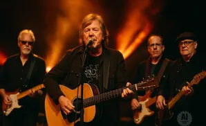 A band performs on stage with warm lighting. The lead singer plays an acoustic guitar.