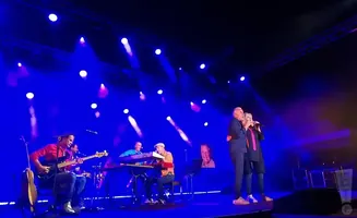 A band performs on a stage with blue lighting. Two singers stand in the foreground.