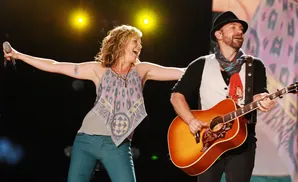 A woman sings into a microphone with her arms outstretched while a man plays an acoustic guitar onstage.