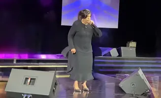 A woman in a black dress sings into a microphone on a stage with purple lighting.