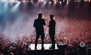 Two singers on stage with a microphone facing a large, cheering crowd, illuminated by stage lights.