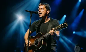 A man sings and plays an acoustic guitar on stage with blue spotlights.