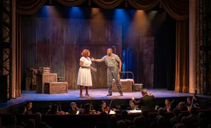 Two actors perform on stage with an orchestra in the foreground.