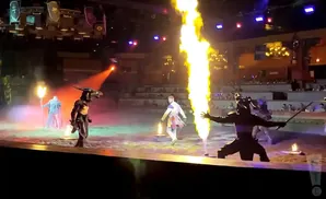 Performers in medieval costumes engage in a sword fight with flames on a stage.