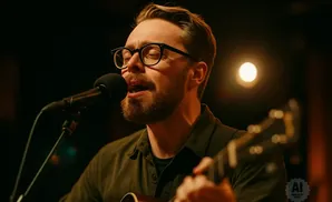 A man with glasses sings into a microphone while playing an acoustic guitar.
