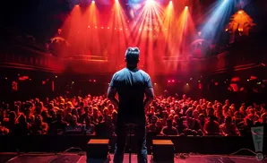 A comedian on stage faces a large audience under bright red stage lights.