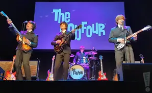 Four musicians dressed as The Beatles play guitars and drums on stage with 