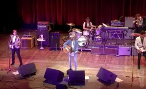 Musicians perform on a stage with red curtains. A guitarist in a sequined jacket is on the left.