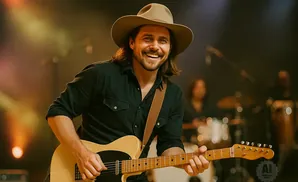 A smiling man in a cowboy hat plays an electric guitar on stage with blurred bandmates in the background.