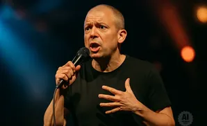A bald man in a black t-shirt holds a microphone and gestures with his chest, speaking animatedly.