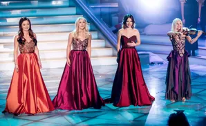 Four women in formal gowns on a stage, one playing the violin.