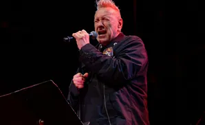 John Lydon sings into a microphone on stage, illuminated by red and blue lights.