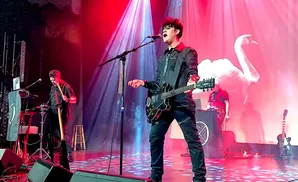 A male singer with dark hair plays a black electric guitar on stage with red and blue lights.