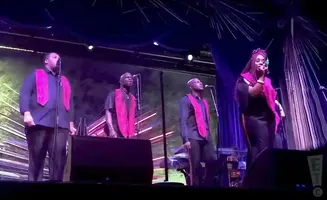 A group of singers on stage wearing pink robes over dark clothing.